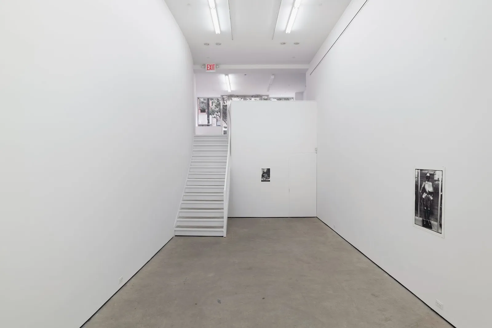 A white gallery room with a staircase leading up to ground level displays two black and white photographs on its walls. The central photo is small. The side photo is larger and depicts a medieval suit of armor.