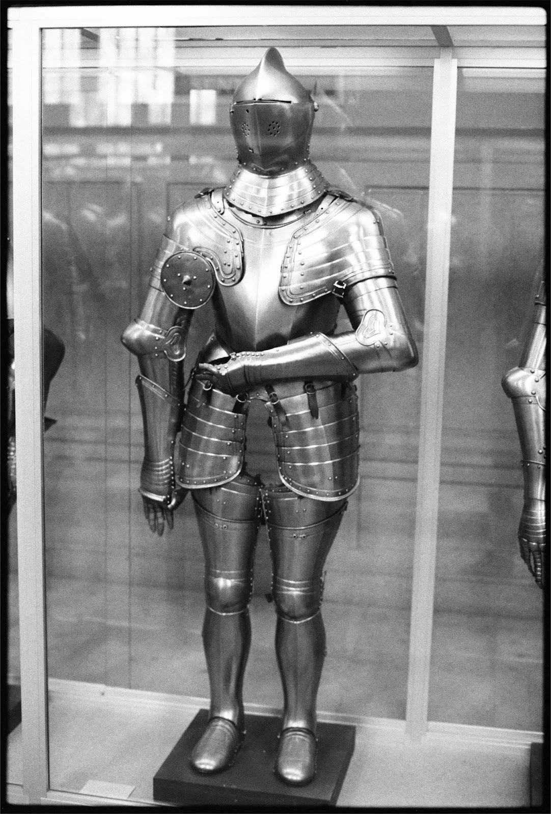 A black-bordered, black and white photograph of medieval armor. The armor's left arm is bent at the elbow and rests across the lower torso. The armor stands behind a display case, with reflections on the glass visible.