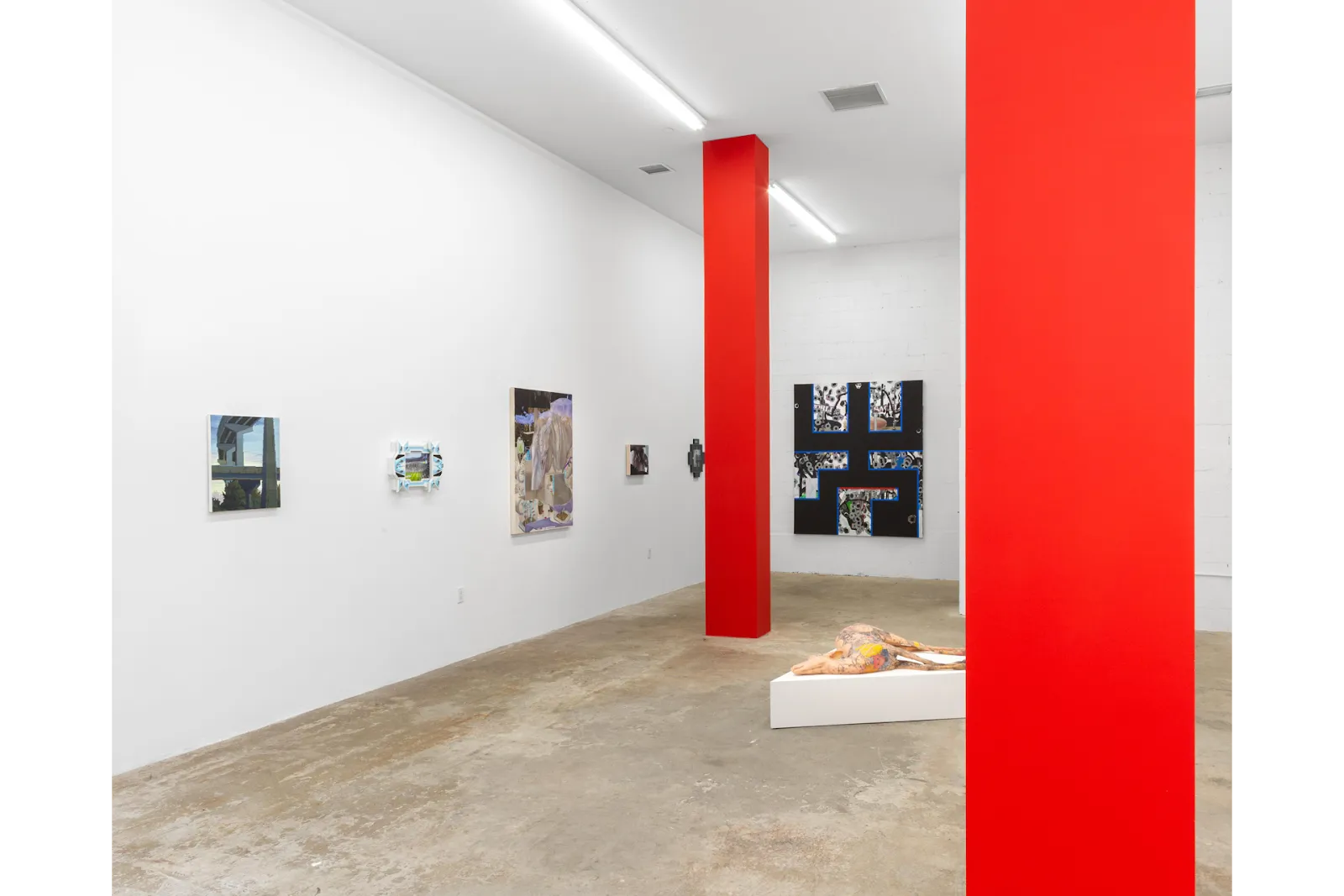 In a white gallery room with bright red supporting columns, various 2D works differing in scale and shape hang on the walls. A central, low triangular plinth features a sculpture in the shape of an animal.