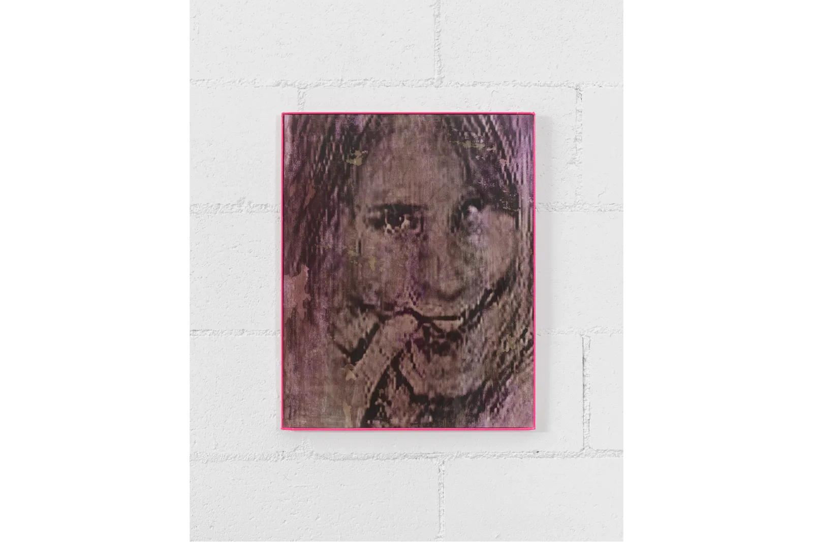 Mounted to a white wall, a thin pink framed 2-D work features a pixelated, smiling face, with eyes staring straight at the viewer.