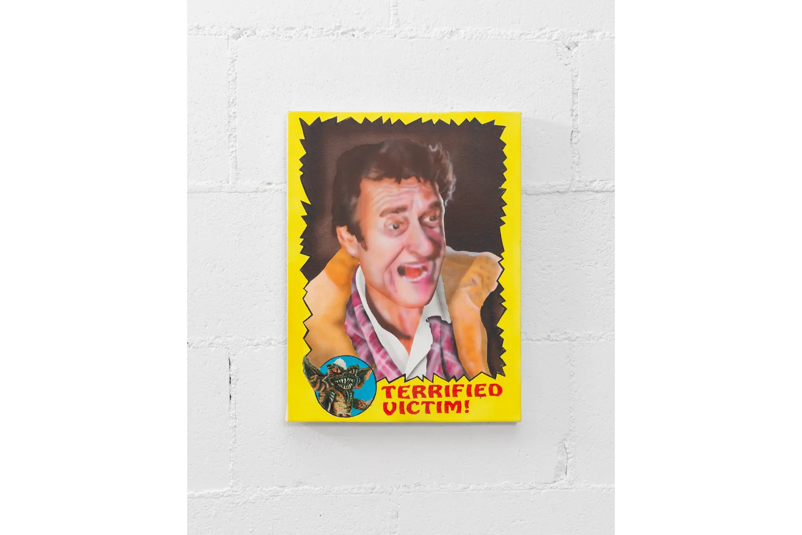 Mounted to a white wall, a graphic work features an airbrushed man with an open-mouthed expression. The border is solid yellow with an irregularly spiked interior. At the bottom, red text reads "terrified victim!" with a gremlin illustration.