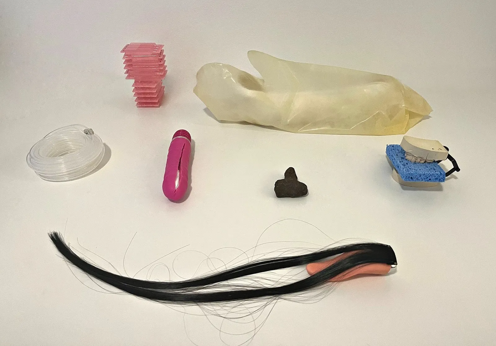 A collection of disparate objects, such as a lock of long black hair, a blue sponge between fake teeth, and clear coiled wire are arranged on a white surface.