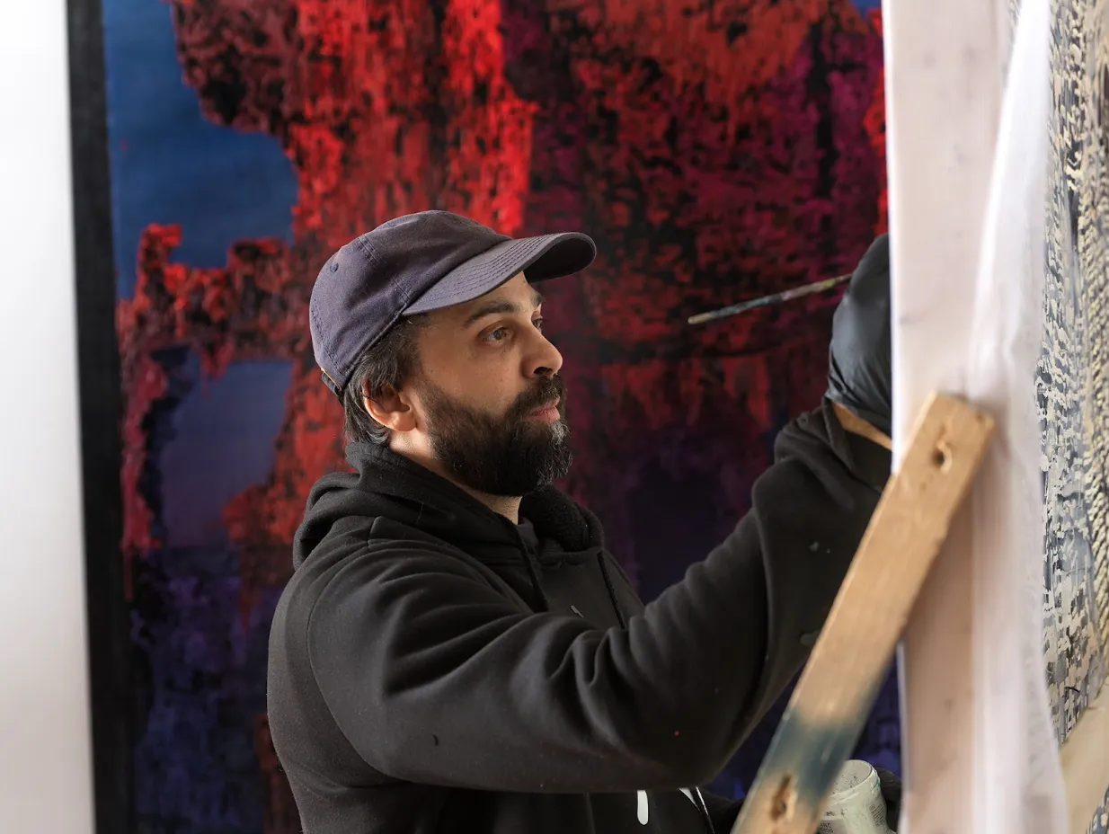 A bearded man in a black sweatshirt and navy blue cap paints on a large canvas.