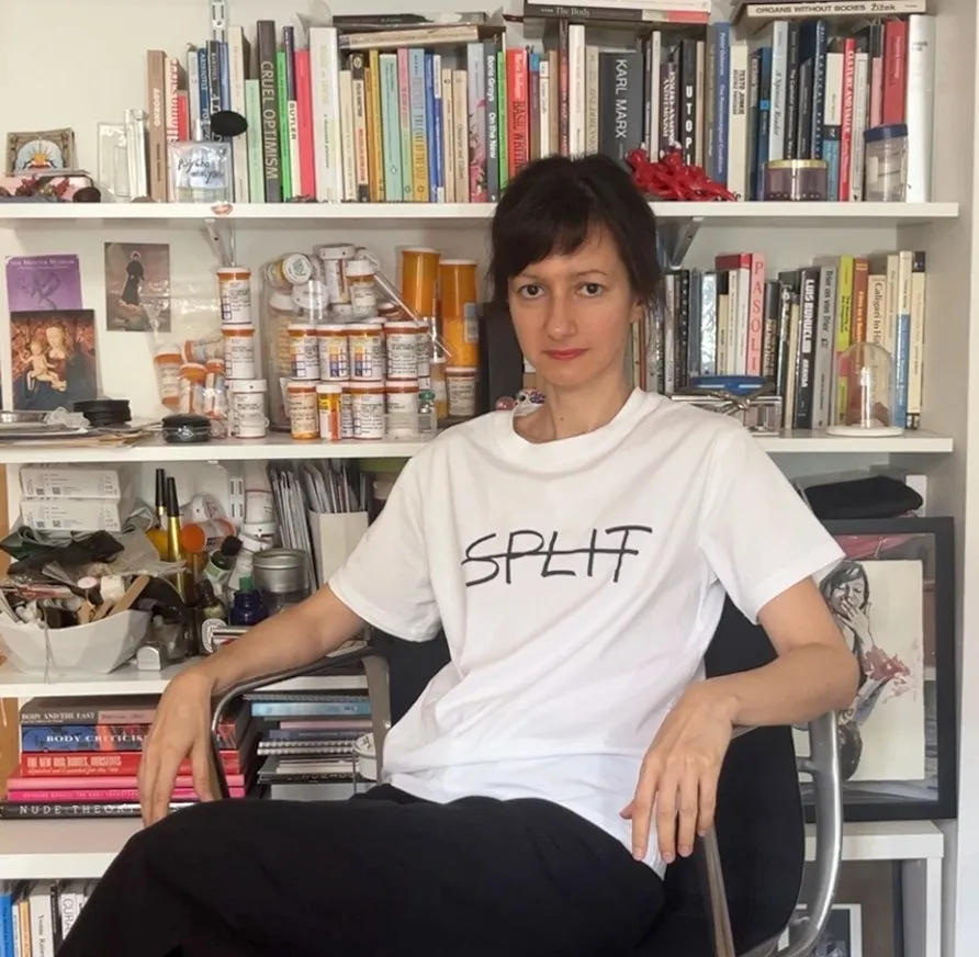 A woman wearing a white t-shirt that reads "SPLIT" sits on a rolling chair in front of a bookcase filled with books and other items.