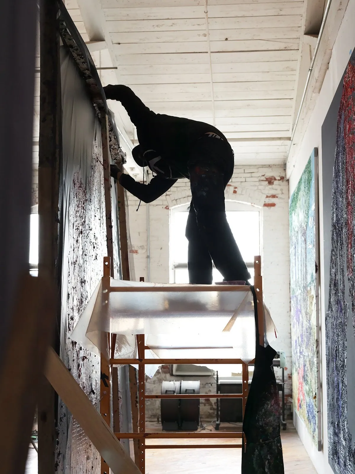 A man's silhouette in front of a large window bends over to paint on a large canvas.