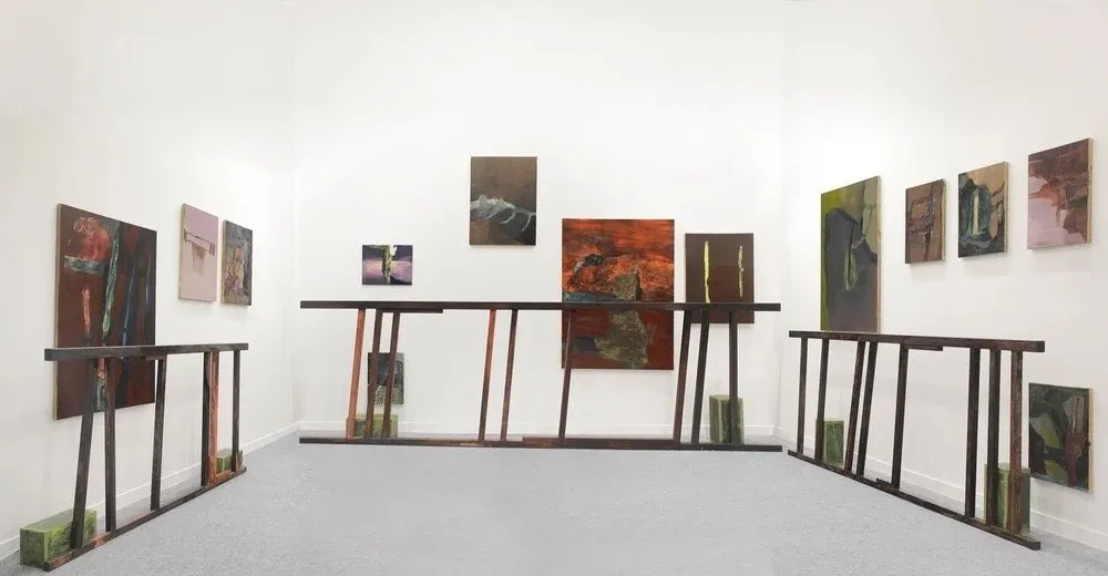 A white gallery room features a number of paintings mounted to each wall and flat wooden palette-like structures standing upright adjacent to each wall.