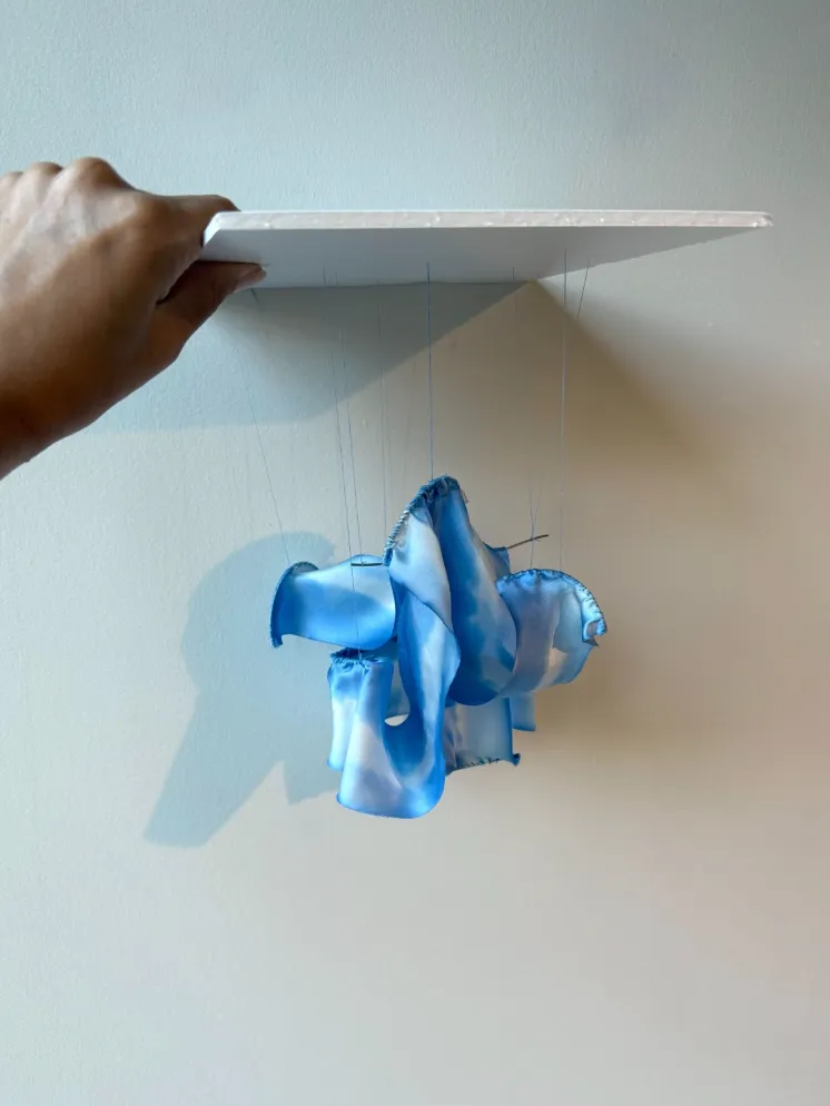 A hand holds up a square of white foam board with threads attached, which support a hanging piece of wavy, folded blue and white cloth.