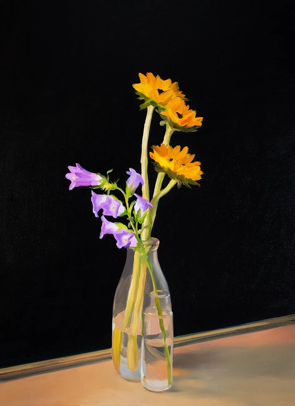 Three sunflowers and purple bellflowers arranged in a glass vase against a black backdrop.