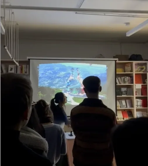 A person stands in front of a projector screen displaying a bright blue landscape, presented to an audience in a room with bookshelves.