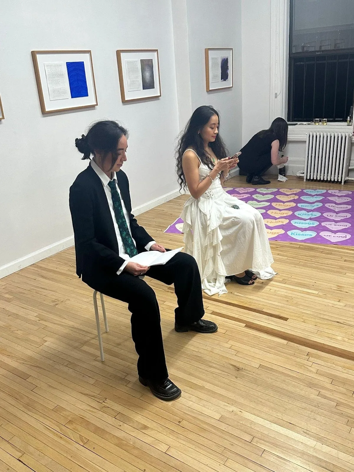 Two people sit on stools in an art gallery; one wears a black suit and tie while reading, and the other wears a white ruffled dress while looking at a phone.