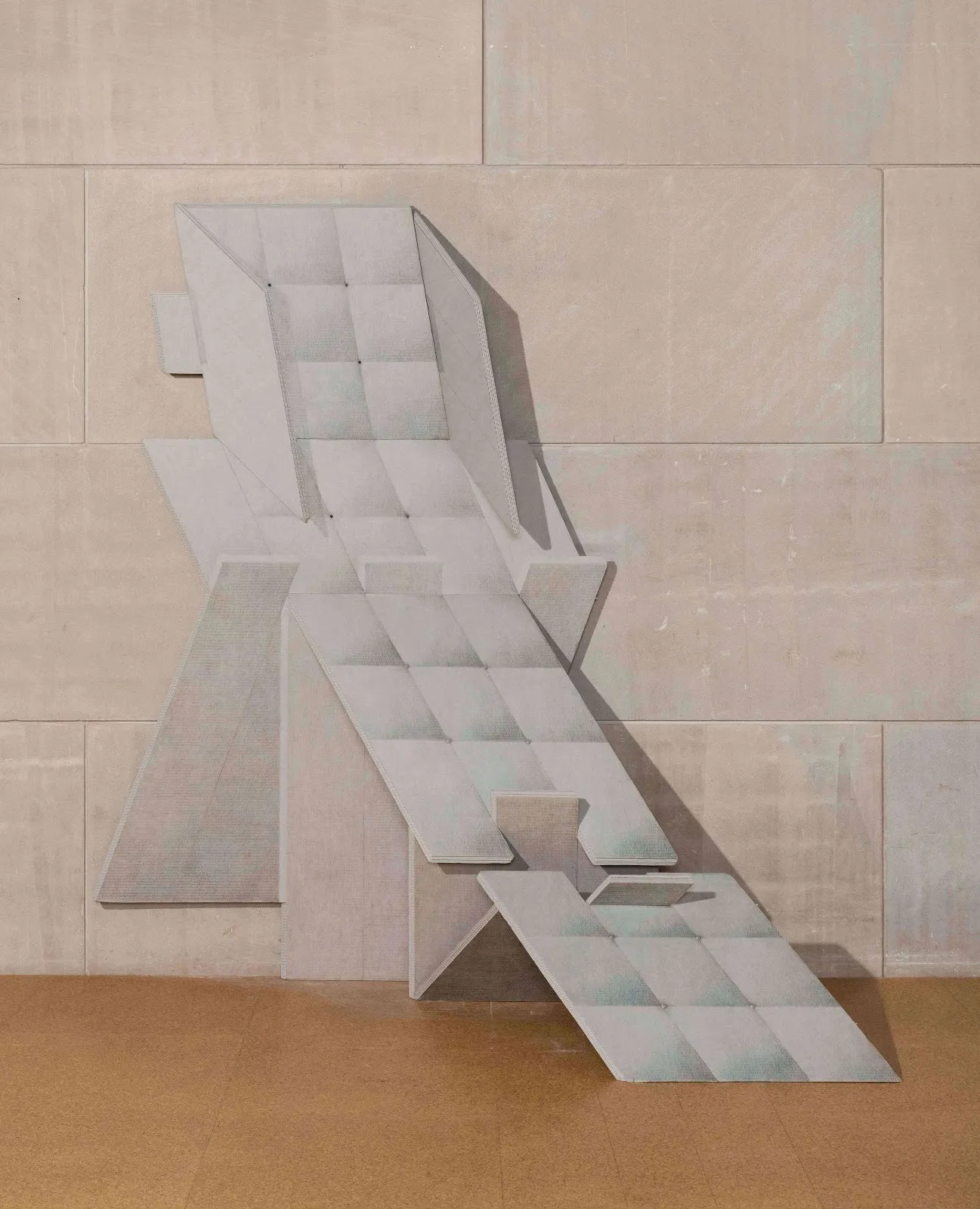A geometric sculpture appears as a structure of folded paper, colored white with numerous folds cascading to form a chair-like shape. The sculpture leans on a wall and the floor.