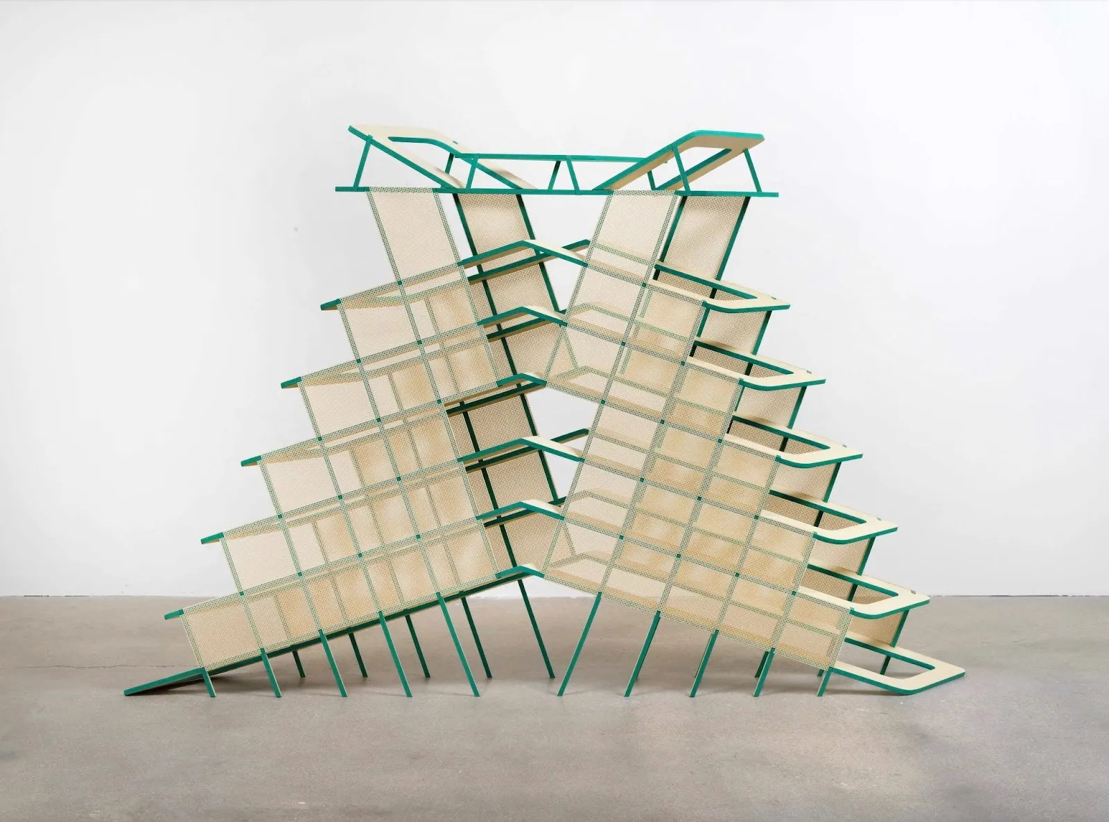 A sculpture is made from geometric, stair-like green metal bars with beige mesh stretched over it. The structure resembles two staircases descending in opposite directions from a center point and tilted away from each other.
