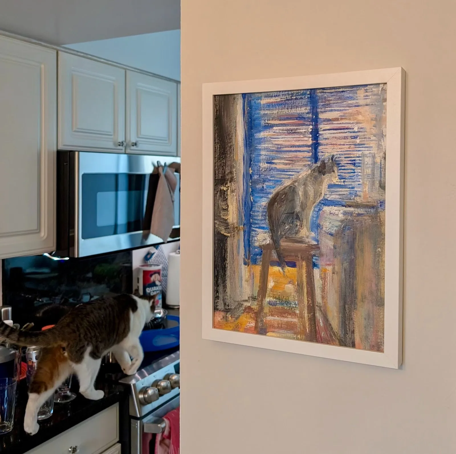 A cat standing on a kitchen stovetop beside glasses and dishes, with a framed painting of a cat on a chair hanging on the adjacent wall.