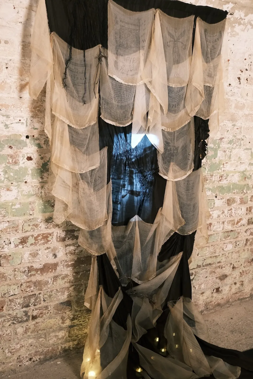 Sheer fabric panels printed with archival text drape over a dark backing, forming a translucent, layered textile installation against a brick wall.