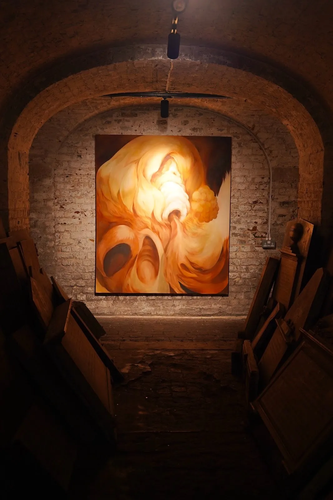 Large abstract painting in warm orange and ochre tones hangs beneath a brick arch in a dimly lit crypt gallery.