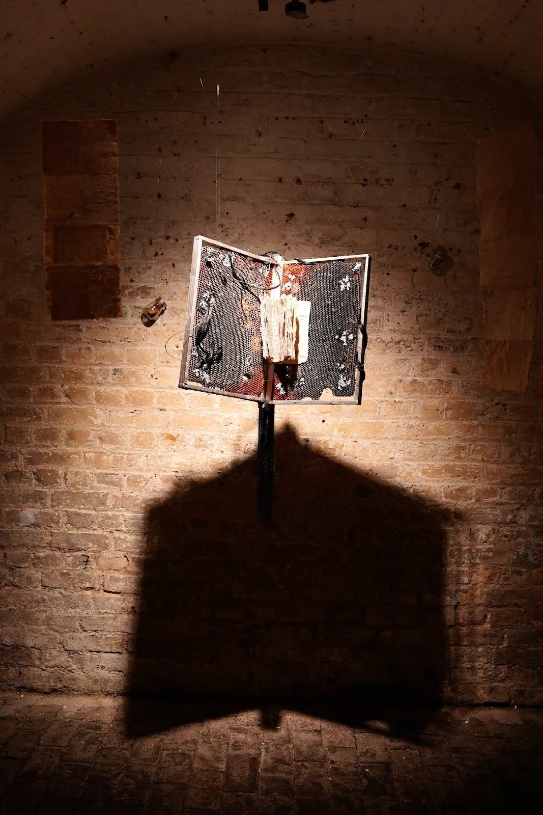 Open book-like sculpture mounted on a stand casts a sharp, wing-shaped shadow onto the brick wall behind it.