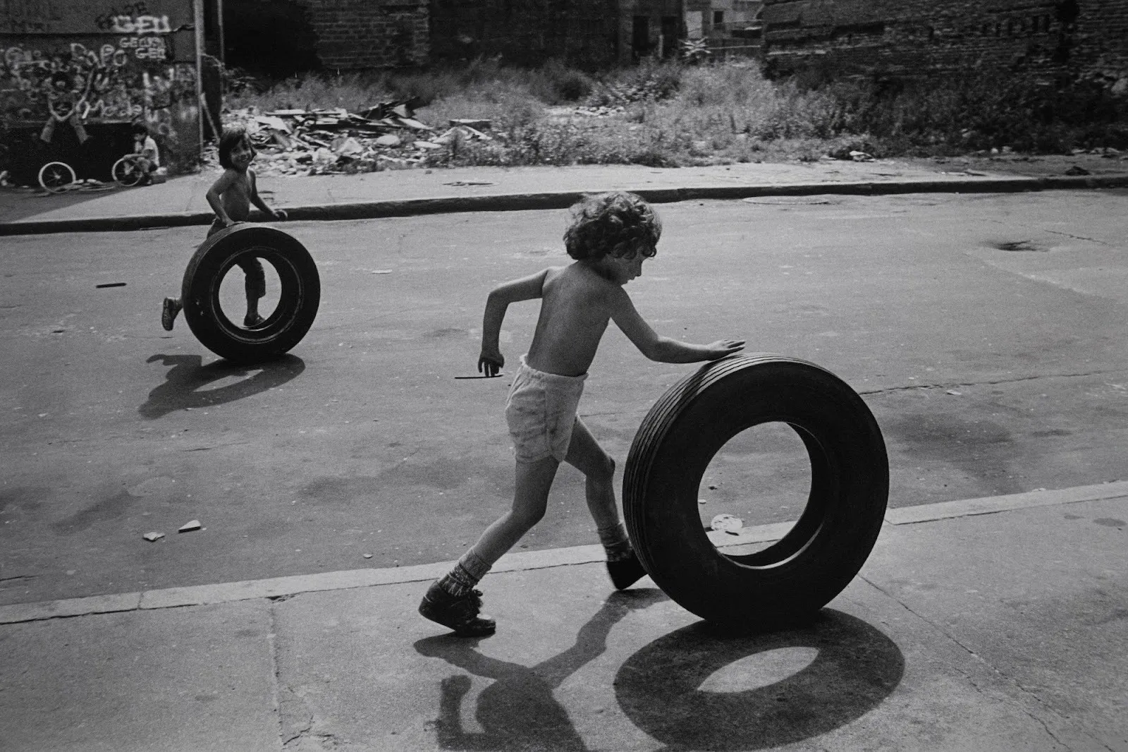 Child pushes a large tire across an empty city street while another follows behind.