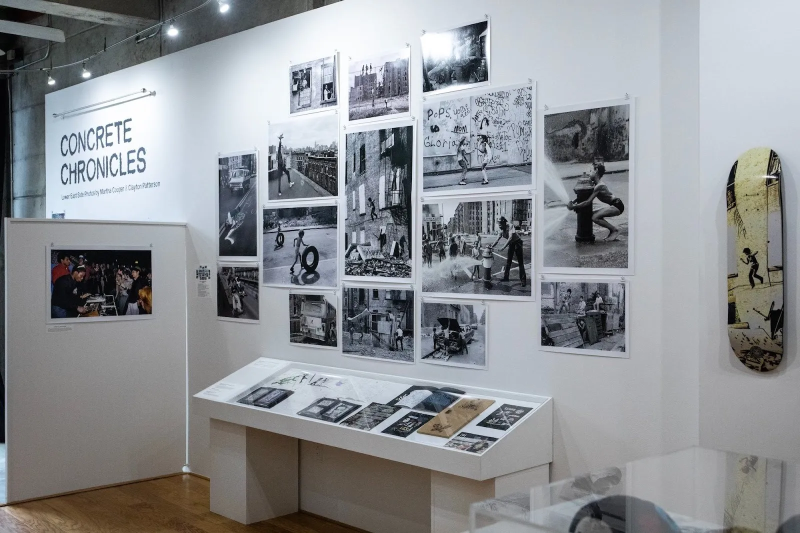 Gallery installation with black-and-white photographs displayed salon-style above a vitrine of archival materials.