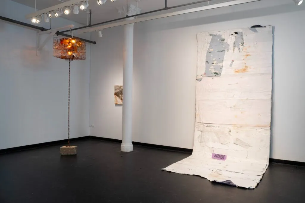 Gallery installation featuring a suspended crab pot with illuminated lights and a large sheet-like wall sculpture.