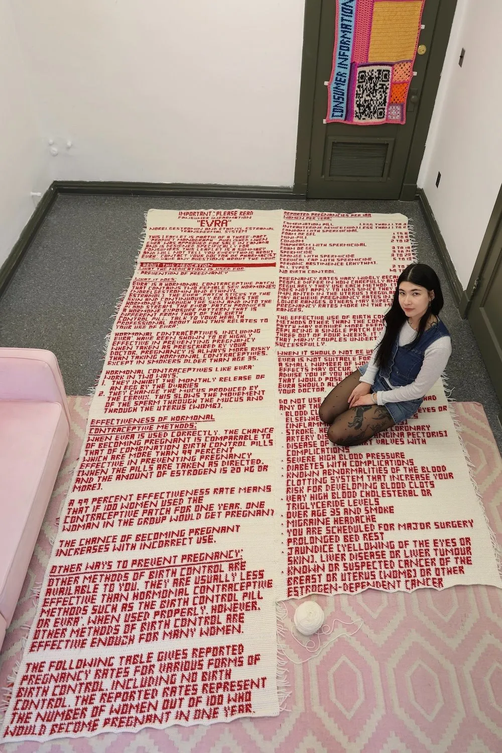 Artist sitting on the floor atop a crocheted red-text textile installation, surrounded by additional textile works hanging on the walls.