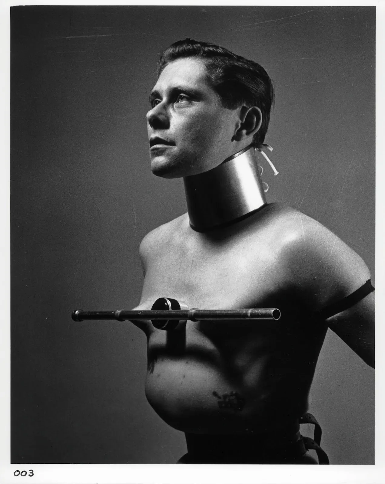 Black-and-white portrait of a bare-chested figure wearing a rigid metal collar and chest apparatus.