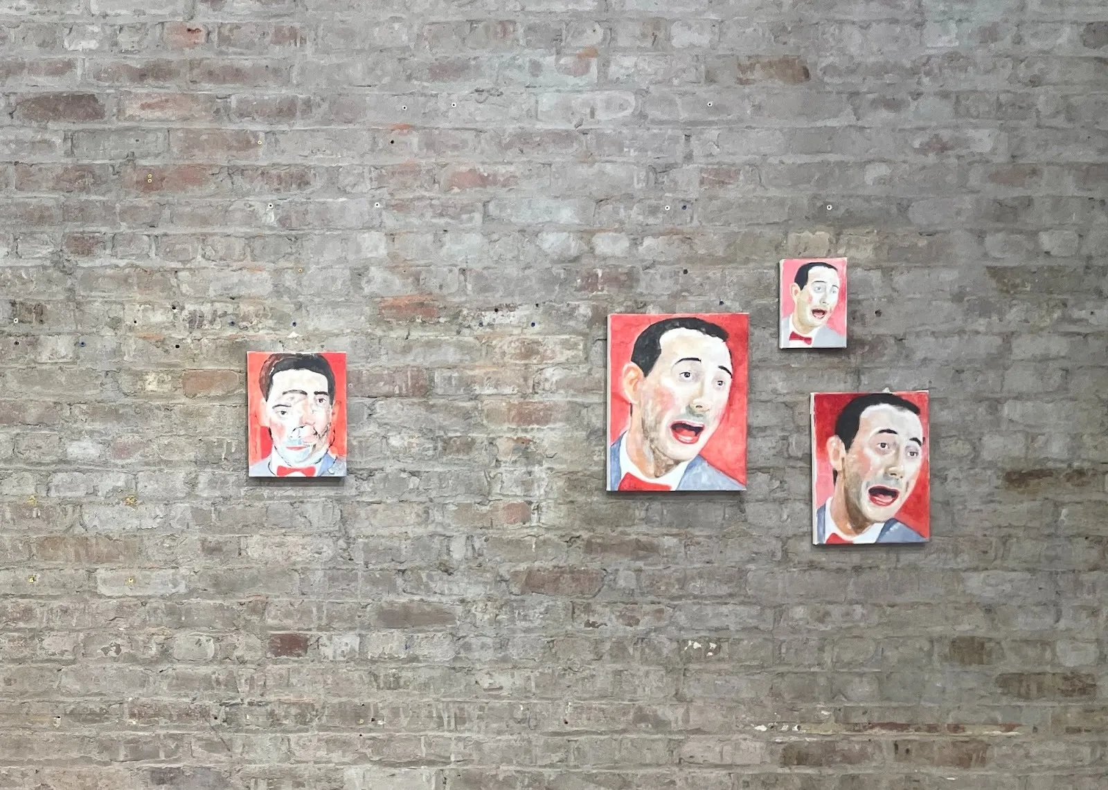 Installation view of four small painted portraits of a man’s expressive face mounted on a brick wall.