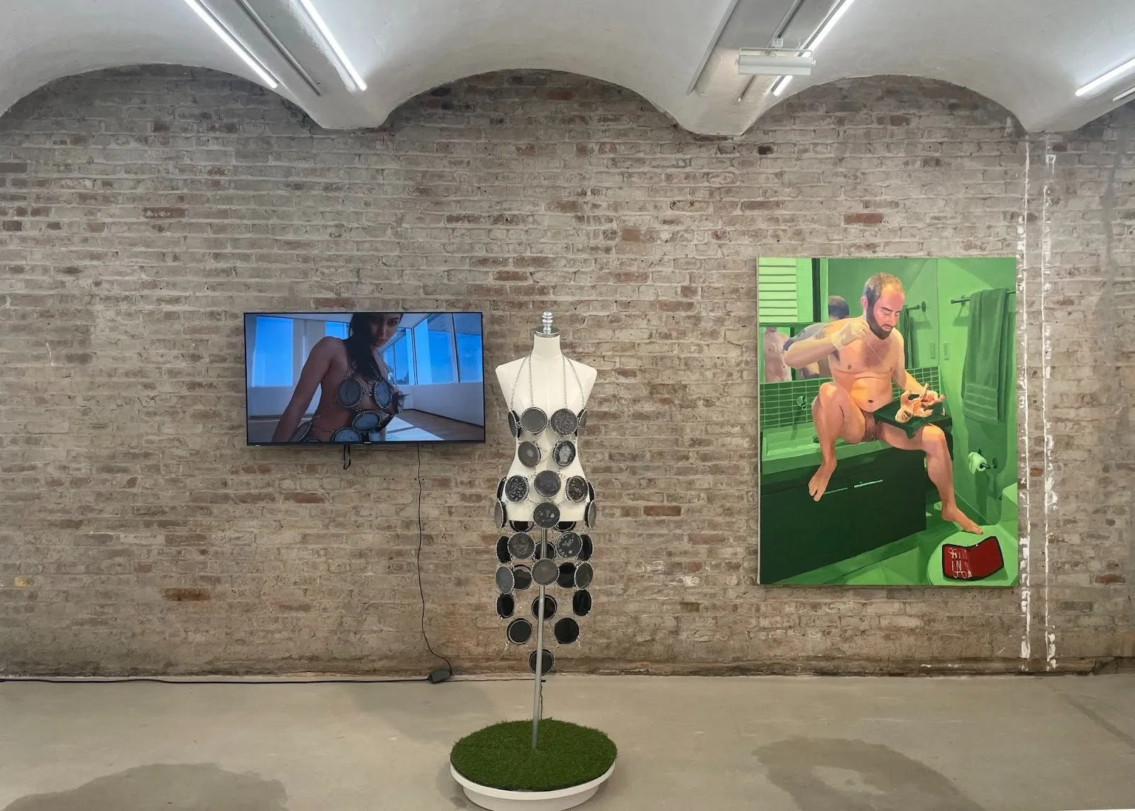 Gallery installation view with a video screen, a mannequin wearing circular metal elements, and a large green-toned painting on a brick wall.