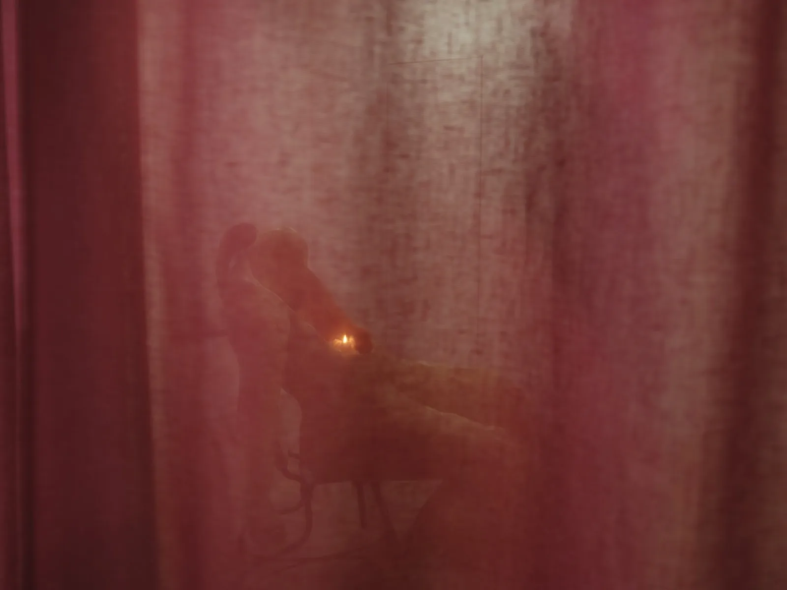 Through a dark red curtain, a sculpture in the shape of a slumped body made of wax and a lit candle can be seen.