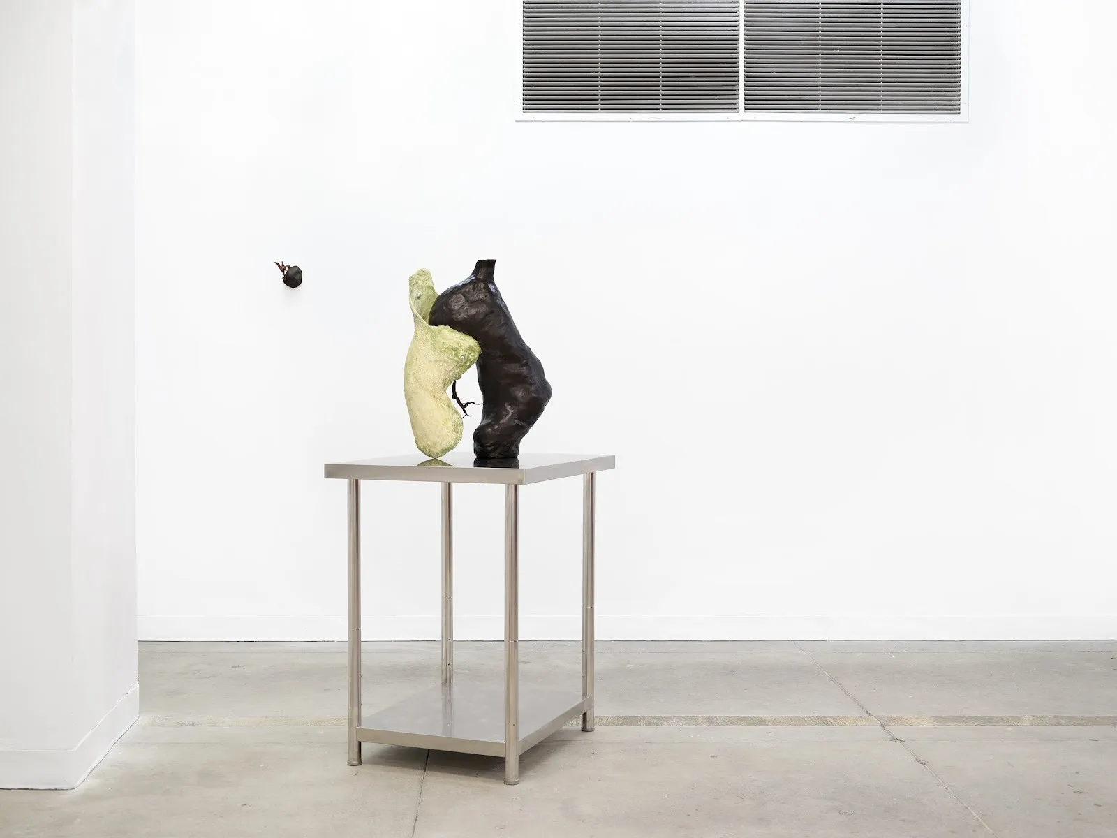 In a white gallery room, a sculpture made of two organic forms coming out of each other, one white and one black, sits on a square steel table.