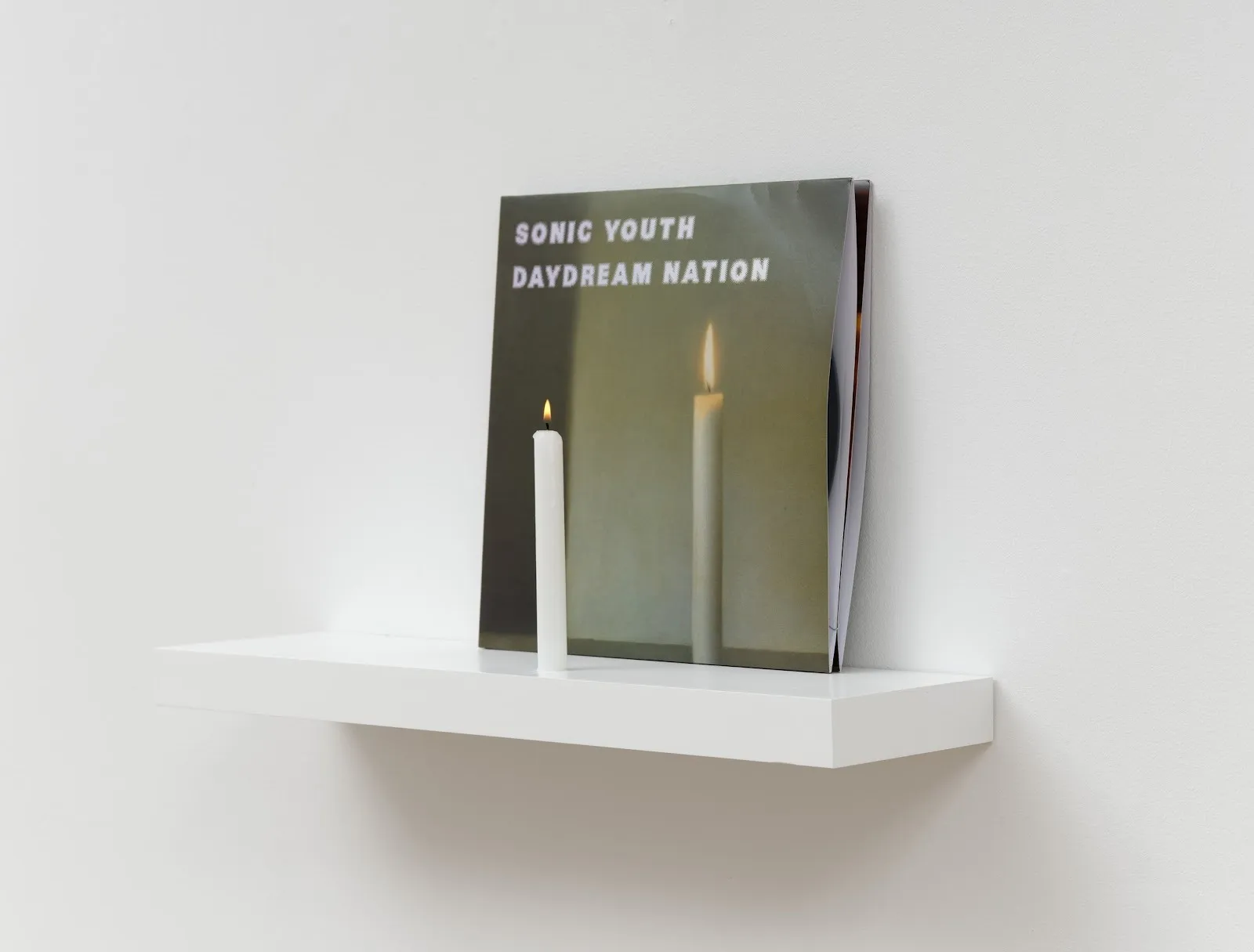 Vinyl record with a lit white candle placed in front, displayed on a white wall shelf.