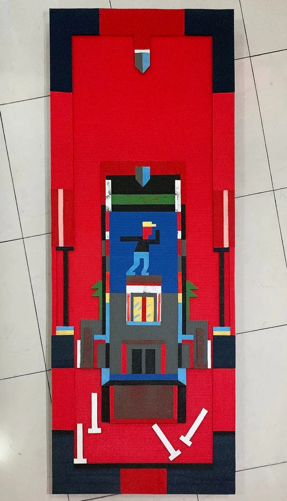 Rectangular textile work in red and black depicting a stylized architectural structure with a small pixelated figure standing atop a platform.