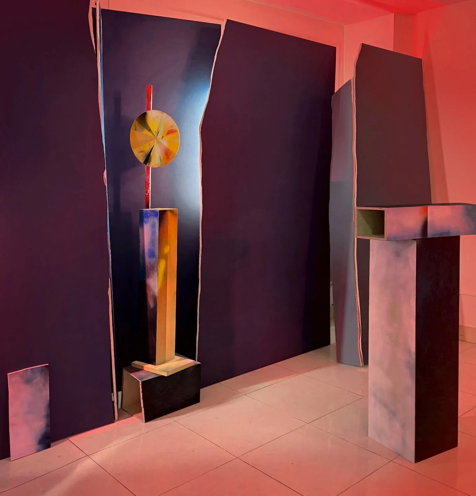 Installation view with freestanding sculptural columns and wall panels, lit in red and blue tones, with a circular geometric element mounted vertically.