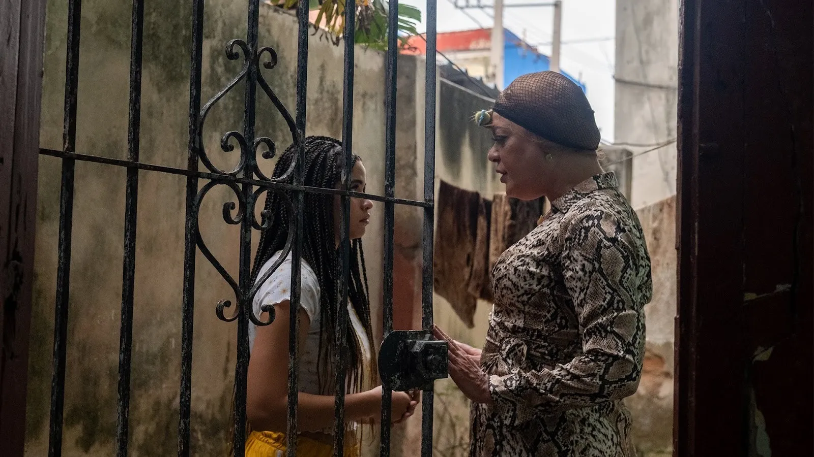 Behind a wrought iron gate door held open by the woman on the left, two women face each other in conversation. They stand in the midst of run-down concrete buildings.