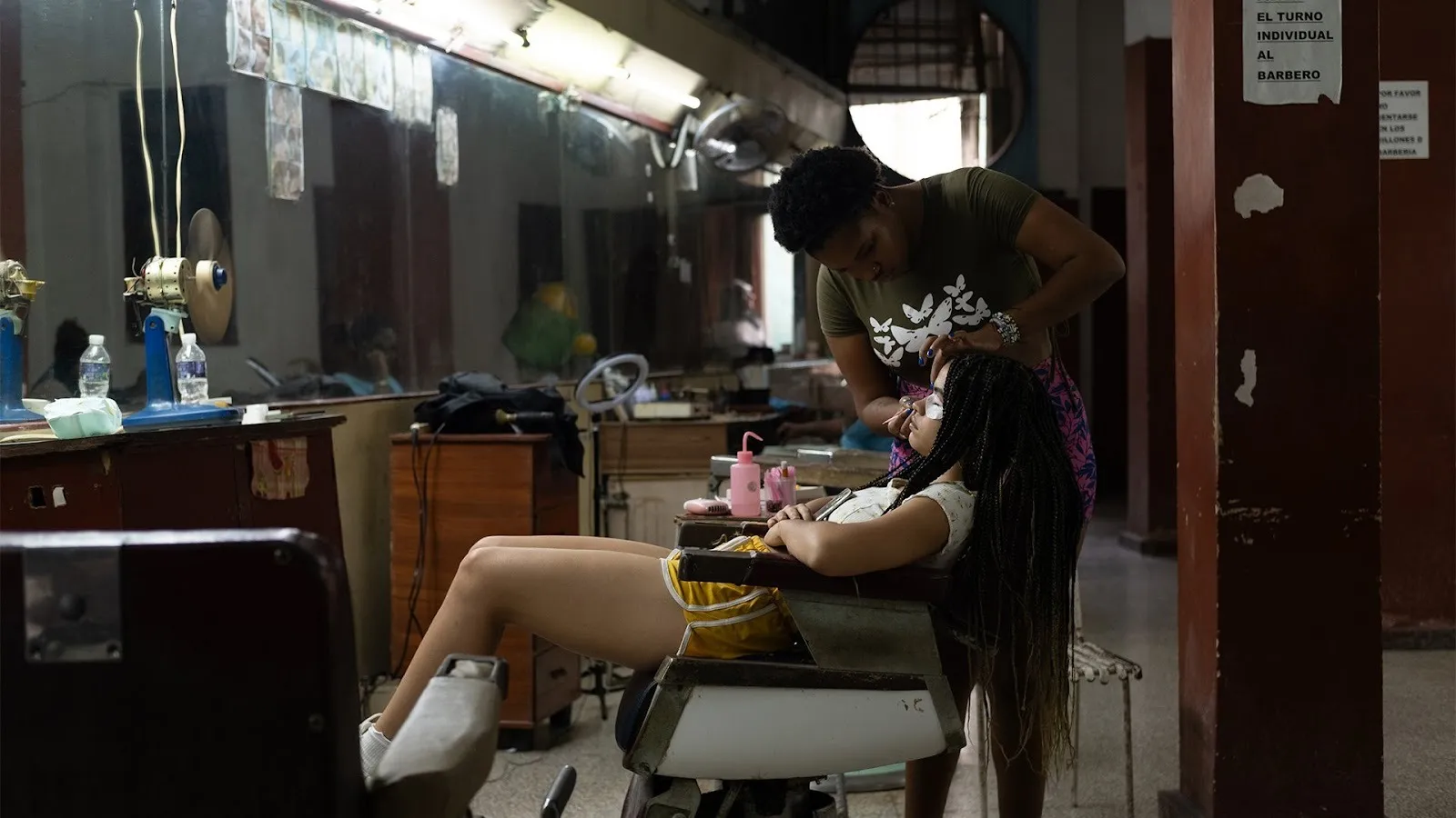 In a dimly-lit hair salon, a girl with long dark hair reclines in a salon chair, eyes closed, while a hairstylist works on her hair. The walls of the salon are fully mirrored from hip-height to ceiling lights.