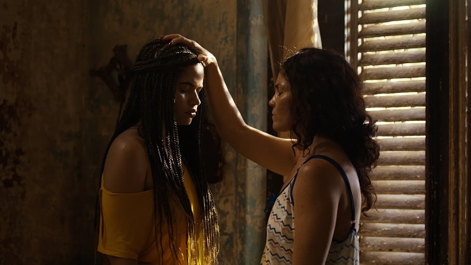 An a dimly lit room with wooden blinds that let in slivers of light, a mother and daughtter face each other, eyes closed. The mother rests her hand on the crown of her daughter's head.