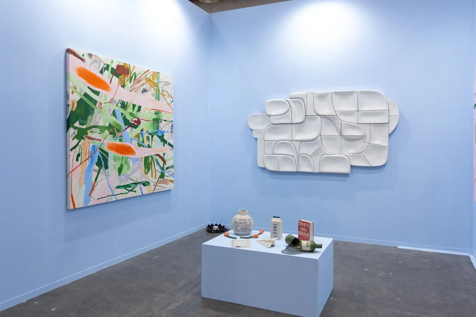 Gallery installation with blue walls, showing an abstract painting, a white modular wall sculpture, and ceramic objects displayed on a central pedestal.