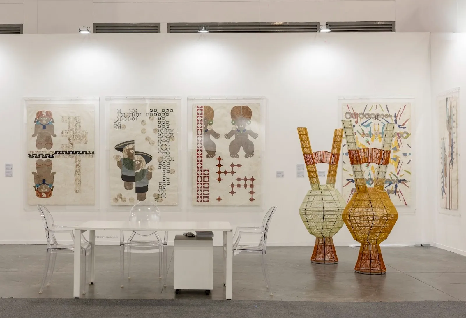 Art fair presentation with framed graphic works, woven sculptural forms, and a white table with clear chairs in a bright booth setting.