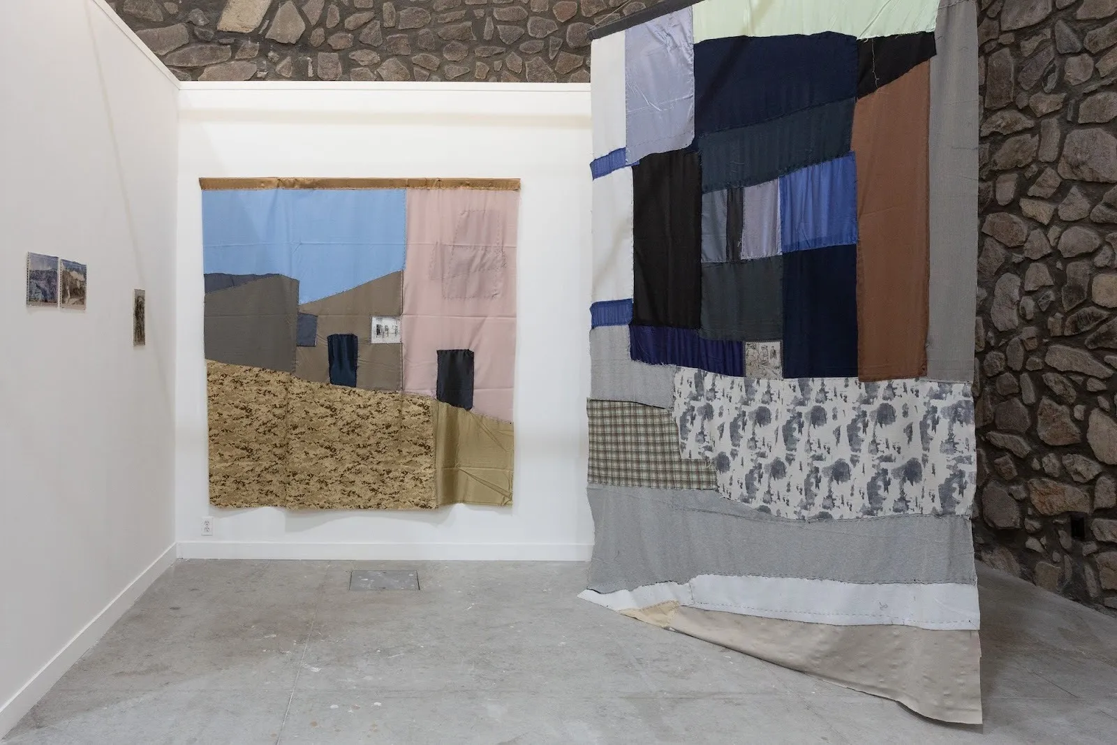 Textile wall works depicting abstracted architectural forms and landscapes, stitched from muted fabric panels in a stone-walled exhibition space.