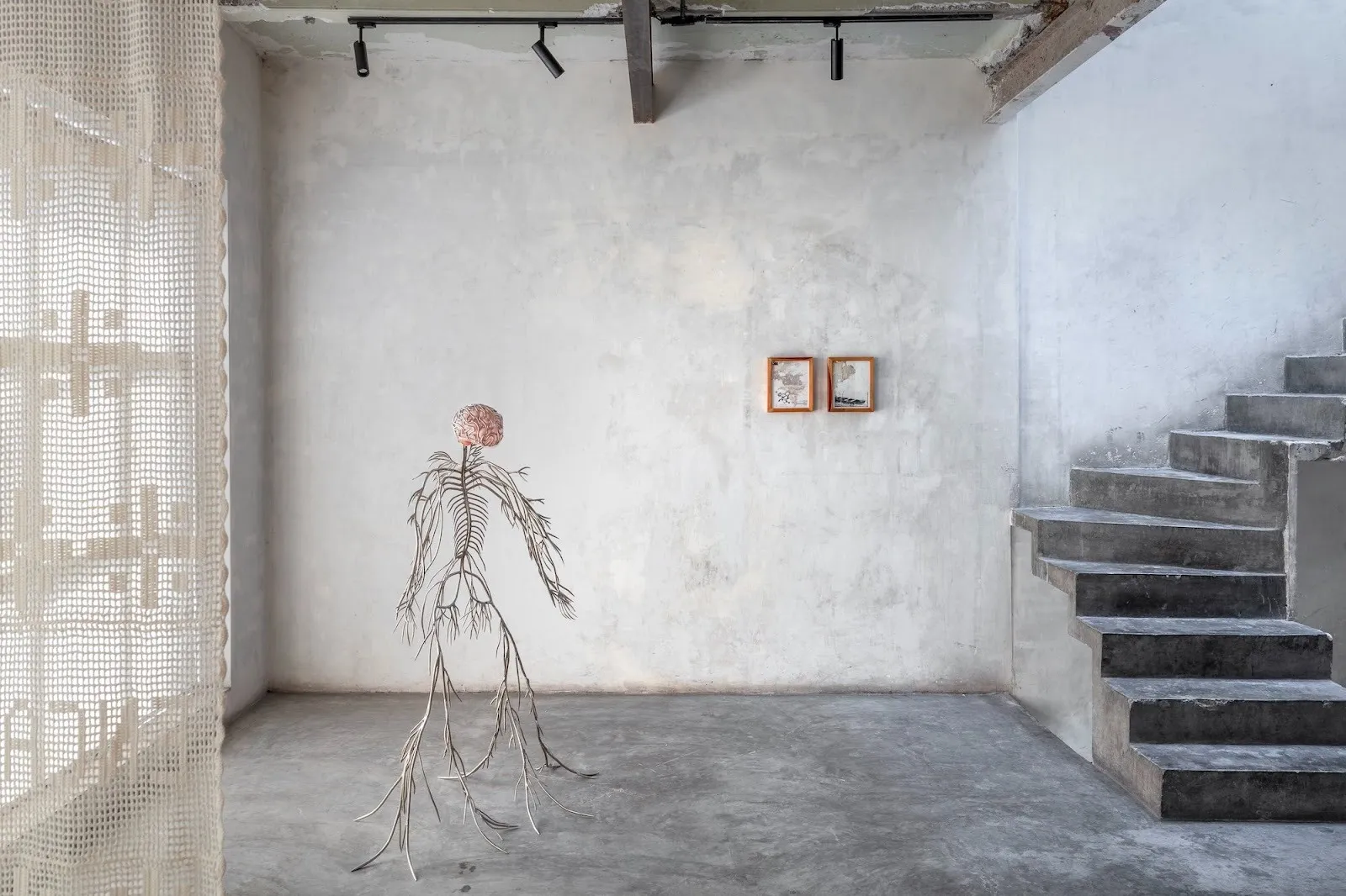 Sculptural figure with branching limbs standing in a raw concrete room, flanked by stairs and framed works on a textured wall.