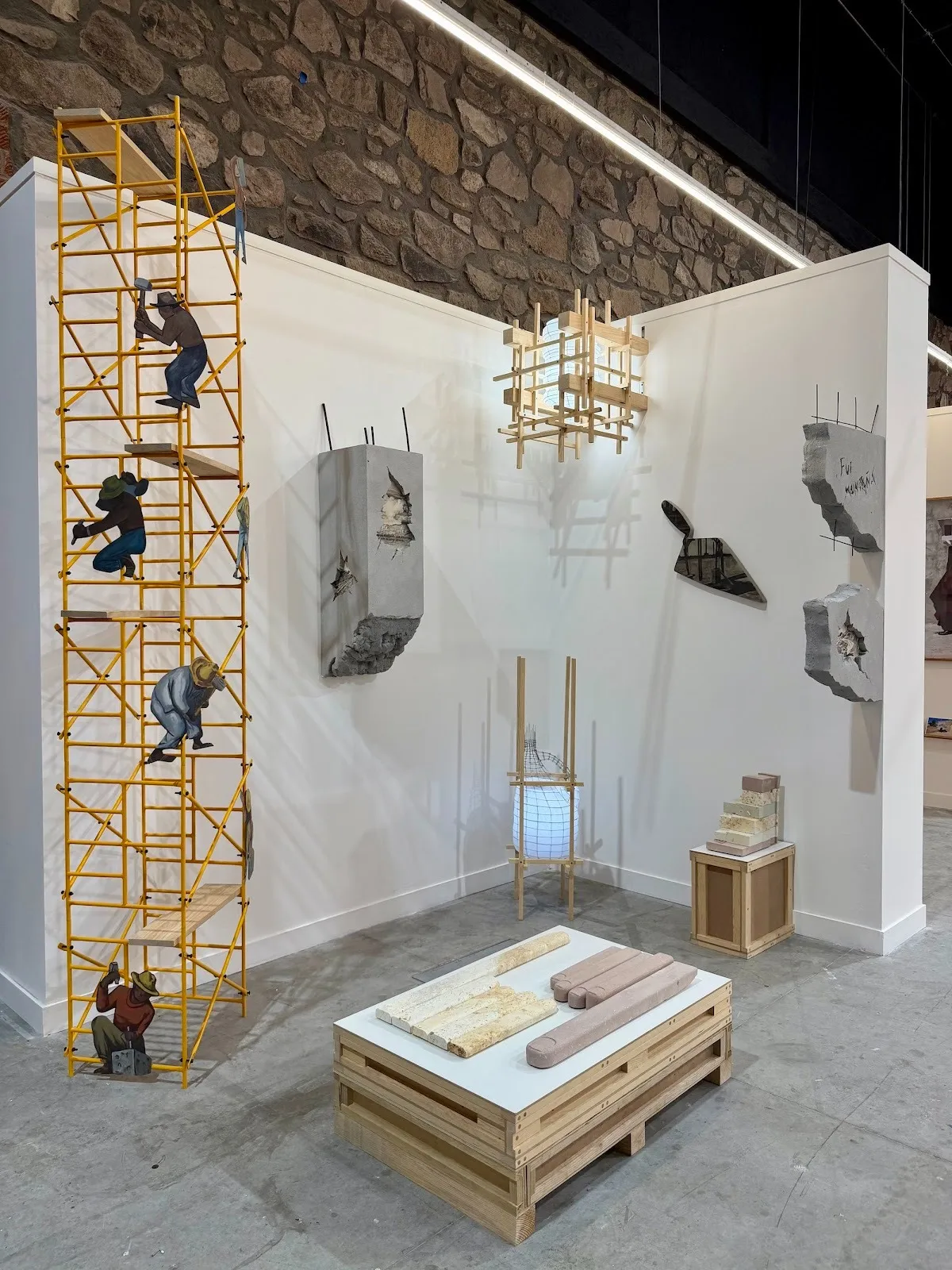 Installation view of mixed-media sculptures, including a yellow scaffold with painted figures, concrete fragments, and wooden structures arranged in a white-walled gallery.