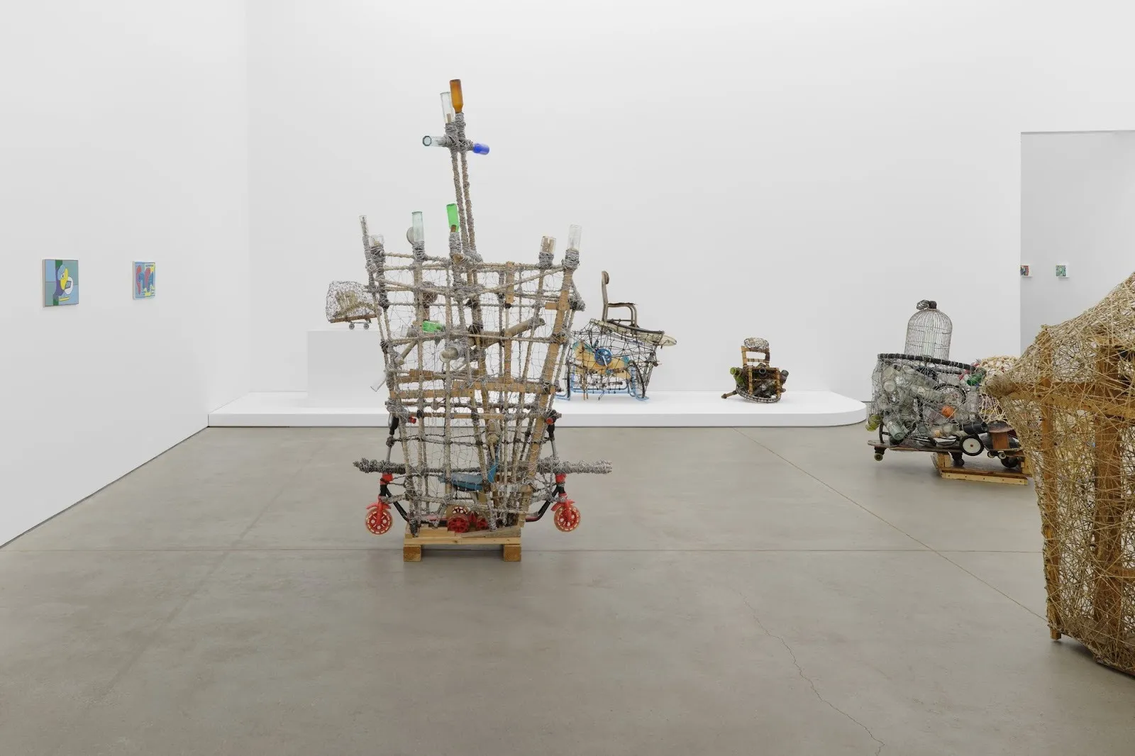 Central gallery view featuring a cross-shaped wire sculpture surrounded by other assemblages on pedestals and low platforms.