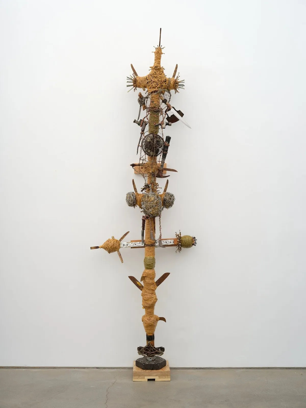 A tall, totem-like assemblage of wrapped wood, wire, tools, and found objects stacked vertically on a small wooden base.