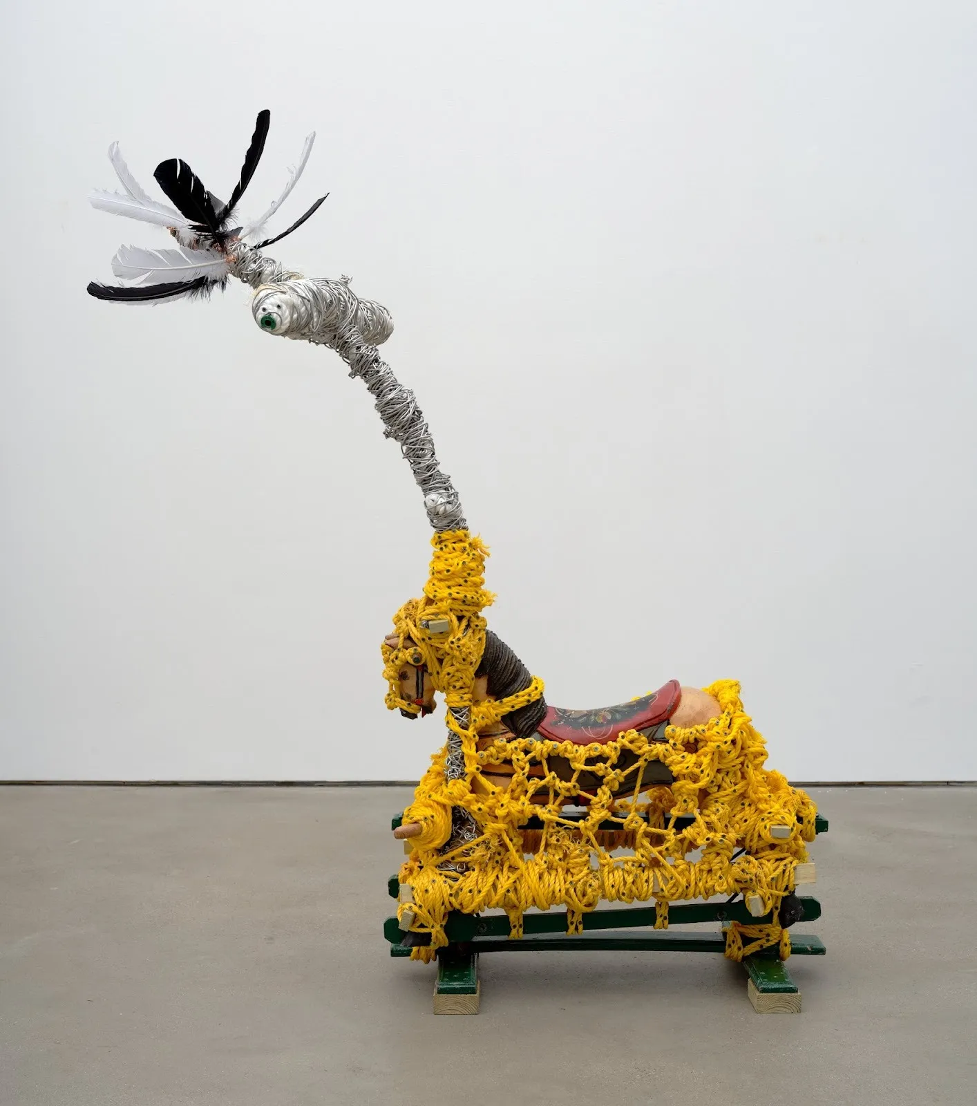 A found-object sculpture wrapped in bright yellow rope, with a curved wire neck topped by feathers, resting on a wooden base.