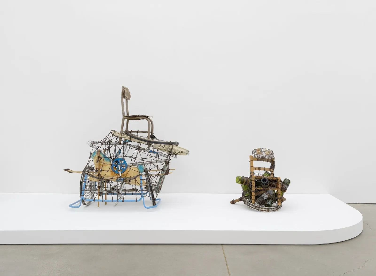Two sculptural objects on a white platform: a wire-framed chair form and a smaller basket-like assemblage filled with bottles.
