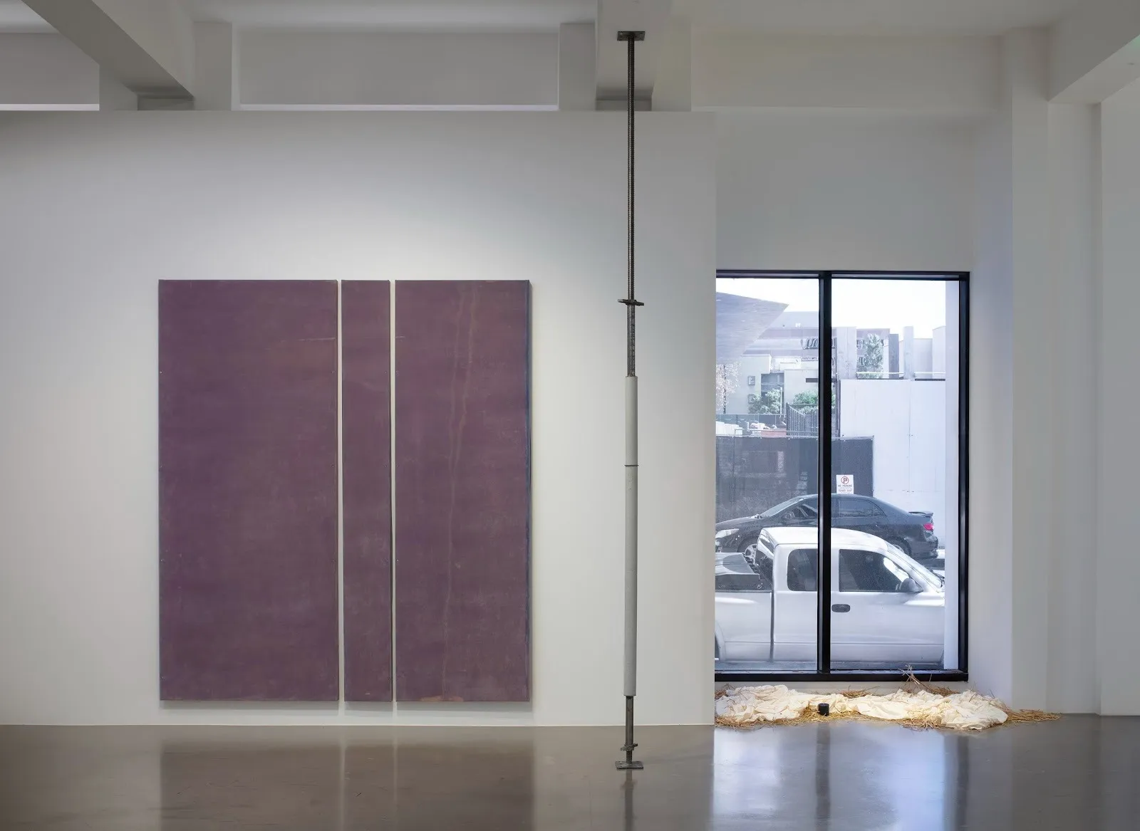 A white gallery room features three works: a large, dark lilac canvas with two vertical white lines, a thin metal pole with two small points of protrusion, and a sculpture on the floor made of a sheet and loose straw.
