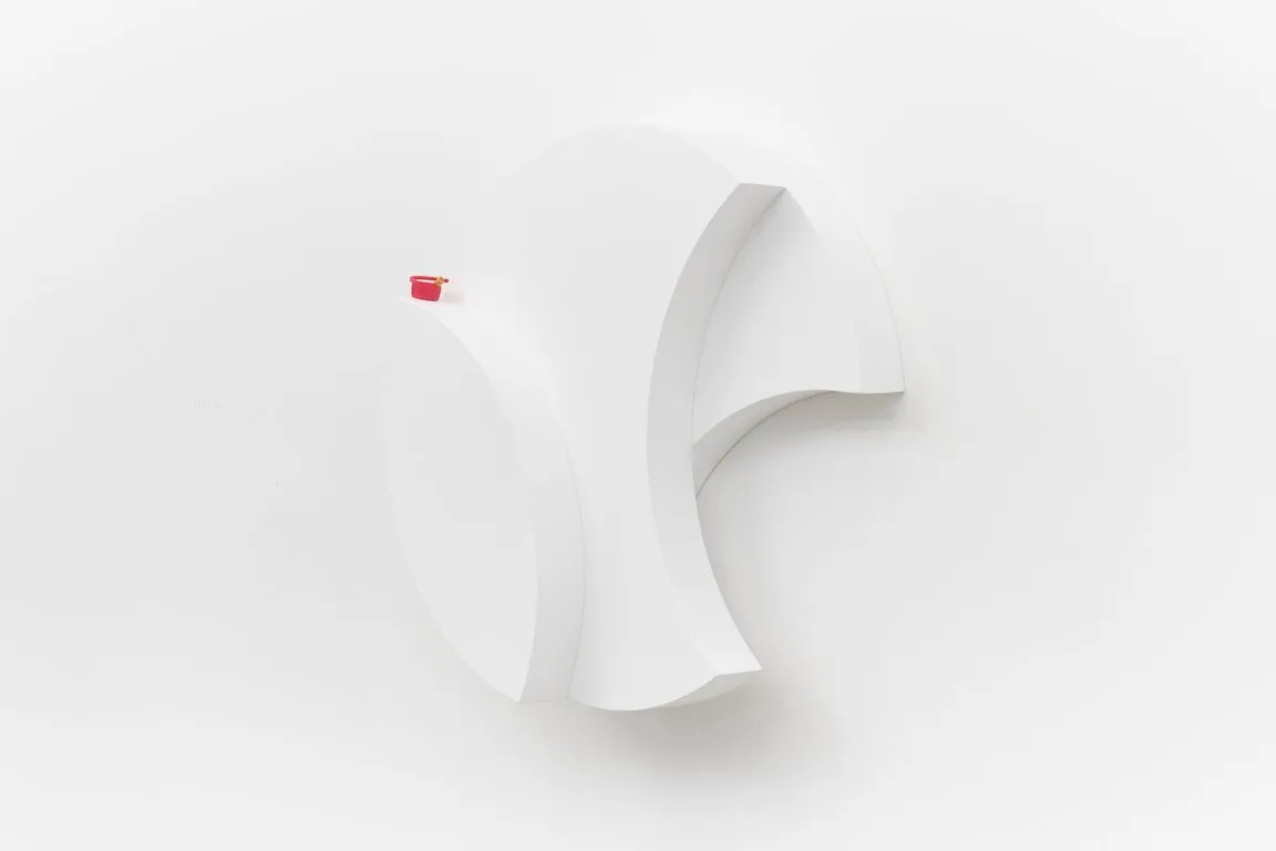 A white sculpture mounted to a white wall consists of overlapping rounded shapes with a small red bottle cap resting on the top of one of the forms.