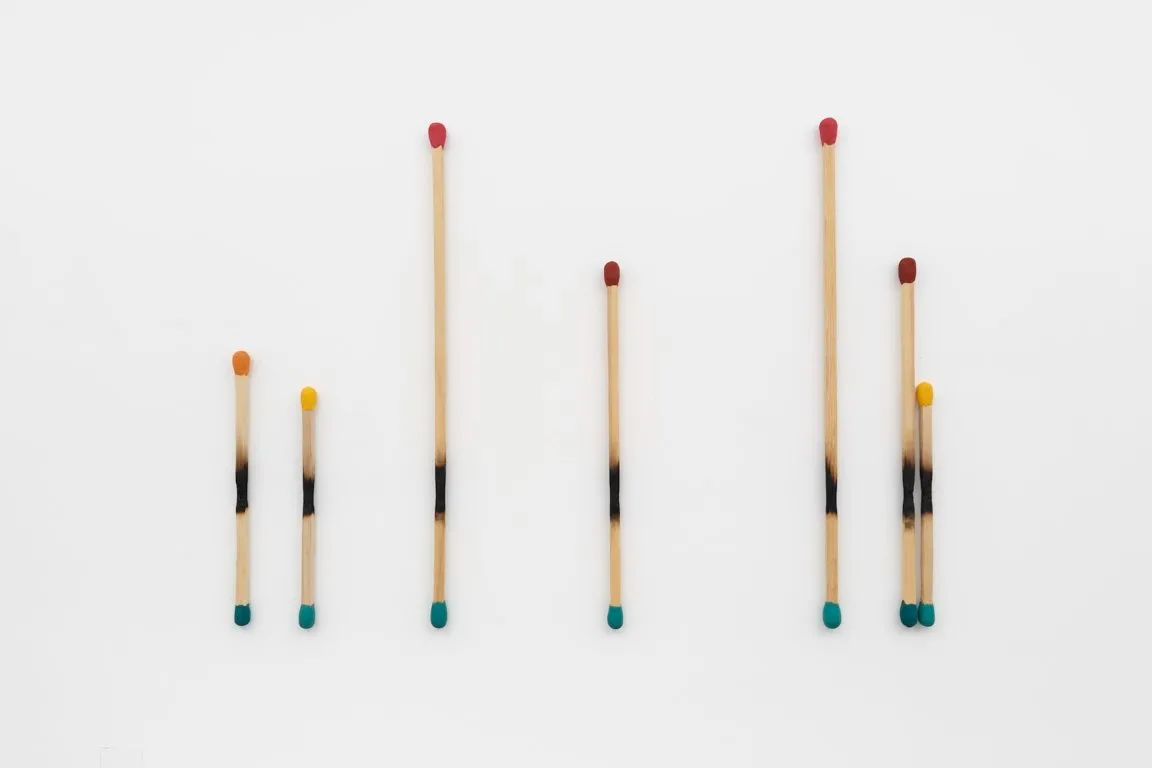 Mounted onto a white wall, a collection of double-headed matchsticks stand at varying lengths and widths apart. Evidence of burning cuts a straight line through the middle of each match.
