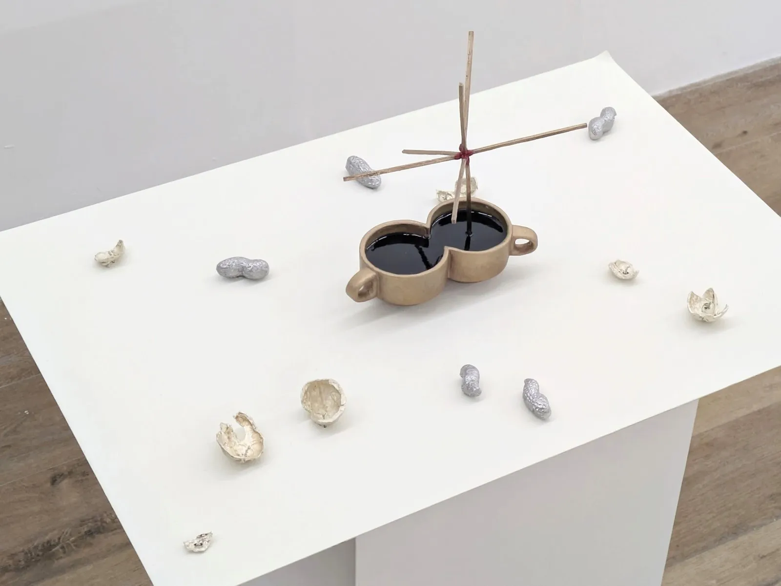 On a white table, a sculpture made of disparate arranged parts consists of silver and gold gilded nut shells and two conjoined metal mugs filled with a dark liquid with a bundle of tied sticks rising from the liquid inside the mugs.