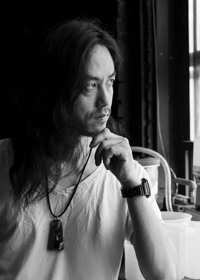 Man with long hair gazing out a window in black and white, resting his hand on his chin in a contemplative pose.