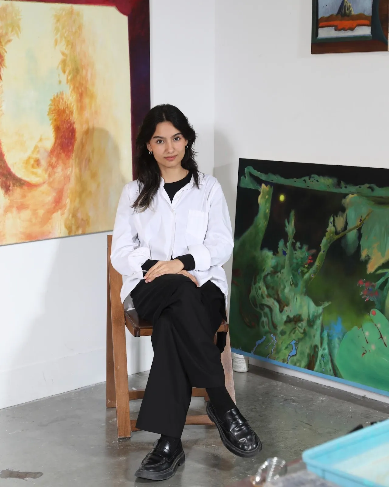 Woman seated on a wooden chair in an art studio, framed by colorful paintings on the wall.